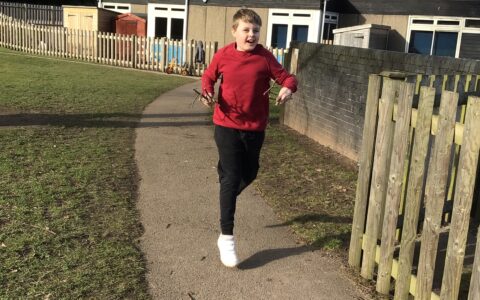 Young boy in red jumper smiling while jogging and carrying sticks